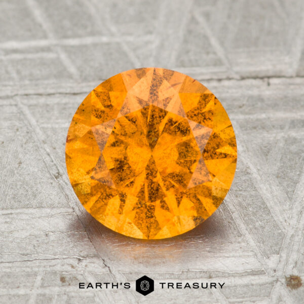 2.10-Carat Spessartine Garnet A 2.10-carat round, faceted Spessartine Garnet gemstone with an orange hue is displayed on a textured gray surface, with the text "Earth's Treasury" underneath.