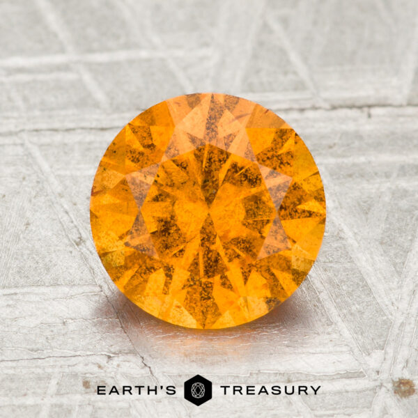 2.10-Carat Spessartine Garnet A 2.10-carat round, faceted Spessartine Garnet gemstone with an orange hue is displayed on a textured gray surface, with the text "Earth's Treasury" underneath.