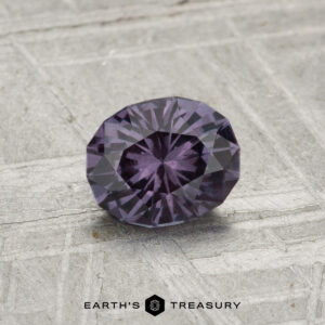 A 0.81-carat color-change garnet, faceted in an oval shape with a deep purple hue, is showcased on a textured gray surface, featuring the "EARTH’S TREASURY" text at the bottom.