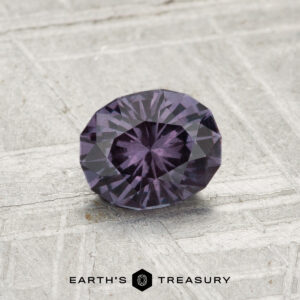 A 0.81-carat color-change garnet, faceted in an oval shape with a deep purple hue, is showcased on a textured gray surface, featuring the "EARTH’S TREASURY" text at the bottom.