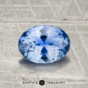 A 1.23-carat Ceylon sapphire, faceted in an oval shape, rests on a textured gray surface with the Earth's Treasury logo and text at the bottom.