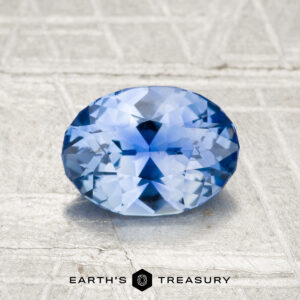 A 1.23-carat Ceylon sapphire, faceted in an oval shape, rests on a textured gray surface with the Earth's Treasury logo and text at the bottom.