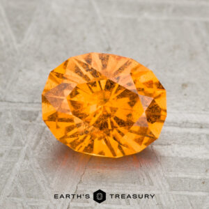 A 2.08-carat Spessartine Garnet gemstone rests on a textured gray surface with the text "EARTH'S TREASURY" at the bottom.