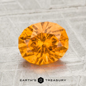 A faceted, oval-cut 2.05-carat Spessartine garnet is displayed on a gray textured surface with the text "EARTH'S TREASURY" below it.