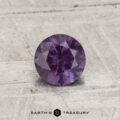 A 0.71-carat color-change garnet, round-cut and purple in hue, is shown on a light textured surface with "EARTH'S TREASURY" at the bottom.