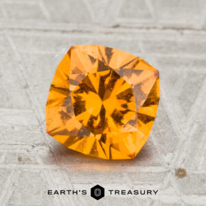 A 1.85-carat Spessartine garnet sits on a textured gray surface, with the "EARTH'S TREASURY" logo and text displayed beneath the gemstone.