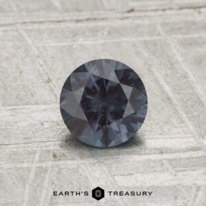 A 0.71-carat color-change garnet, round and faceted, rests on a textured, light surface with "EARTH'S TREASURY" displayed below.