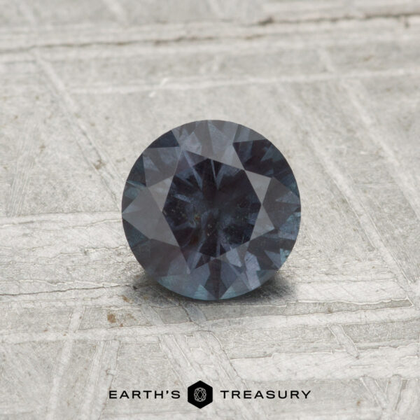 A 0.71-carat color-change garnet, round and faceted, rests on a textured, light surface with "EARTH'S TREASURY" displayed below.
