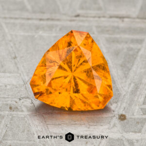 A 1.82-carat Spessartine garnet, faceted in a triangular shape, sits on a textured gray surface with the "Earth's Treasury" logo at the bottom.
