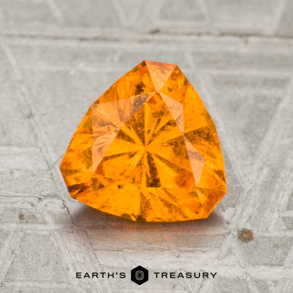 1.82-Carat Spessartine Garnet A 1.82-carat Spessartine garnet, faceted in a triangular shape, sits on a textured gray surface with the "Earth's Treasury" logo at the bottom.