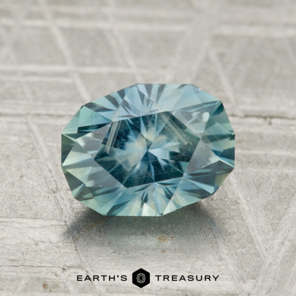 1.99-Carat Teal Montana Sapphire A 1.99-carat Montana sapphire, faceted in an oval cut with blue-green hues, rests on a textured gray surface. Below is the text “EARTH’S TREASURY.”.