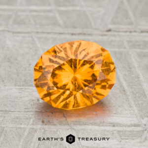 A 1.69-carat faceted oval Spessartine Garnet with vibrant orange color is displayed on a textured gray surface, featuring the text "EARTH'S TREASURY" at the bottom.