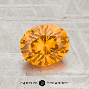 A 1.69-carat faceted oval Spessartine Garnet with vibrant orange color is displayed on a textured gray surface, featuring the text "EARTH'S TREASURY" at the bottom.