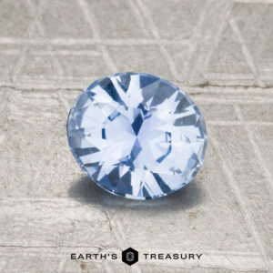 A 1.12-carat Ceylon Sapphire, round and faceted in light blue, rests on a textured gray surface with "EARTH'S TREASURY" displayed at the bottom.