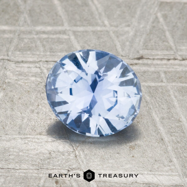1.12-Carat Sky Blue Ceylon Sapphire A 1.12-carat Ceylon Sapphire, round and faceted in light blue, rests on a textured gray surface with "EARTH'S TREASURY" displayed at the bottom.