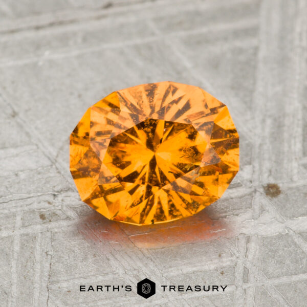 1.66-Carat Spessartine Garnet A 1.66-carat spessartine garnet, round brilliant-cut, is shown on a textured metallic surface with "EARTH'S TREASURY" beneath it.