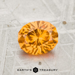 A 1.55-carat round brilliant-cut light orange Spessartine Garnet is displayed on a textured gray surface, with "EARTH'S TREASURY" written below the gemstone.