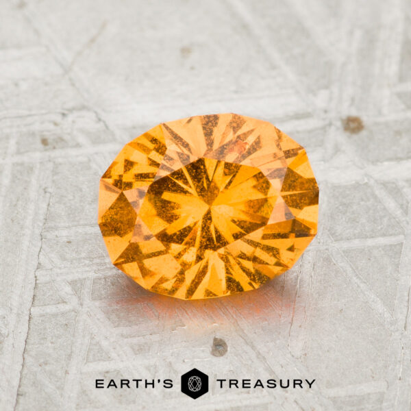 1.55-Carat Spessartine Garnet A 1.55-carat round brilliant-cut light orange Spessartine Garnet is displayed on a textured gray surface, with "EARTH'S TREASURY" written below the gemstone.
