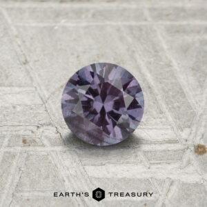 A 0.54-carat color-change garnet, round and faceted, is shown on a textured gray surface with the "EARTH'S TREASURY" logo at the bottom.