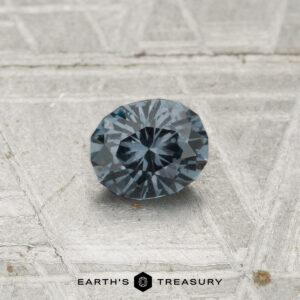 A 0.44-carat color-change garnet, round and faceted in dark gray, is shown on a textured light gray surface with "EARTH'S TREASURY" text at the bottom.