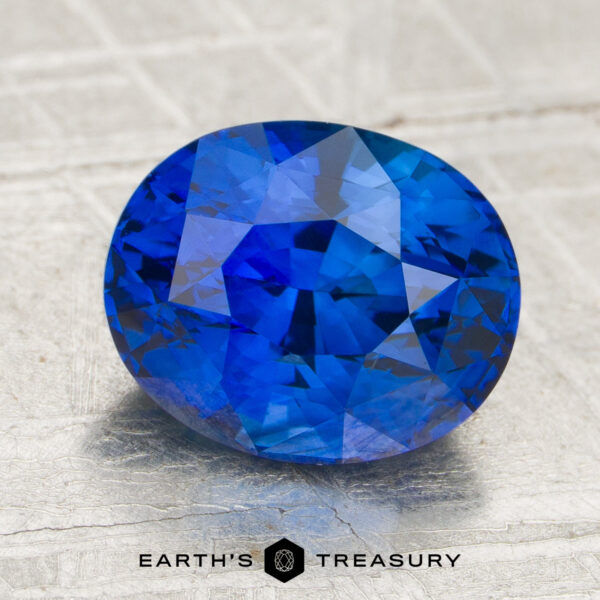 2.63-Carat Sapphire (Heated) A 2.63-carat royal blue Ceylon sapphire (heated) in an oval faceted cut rests on a textured gray surface, with the "Earth's Treasury" logo and text displayed beneath it.