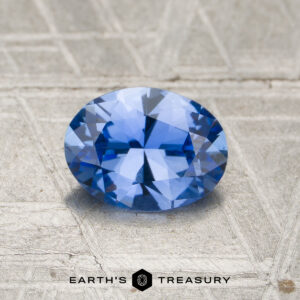 A 0.97-carat unheated Cornflower Blue Ceylon Sapphire, faceted oval, shown on a textured metallic surface with "EARTH'S TREASURY" and logo below.
