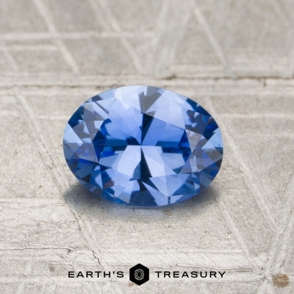 0.97-Carat Cornflower Blue Ceylon Sapphire (Unheated) A 0.97-carat unheated Cornflower Blue Ceylon Sapphire, faceted oval, shown on a textured metallic surface with "EARTH'S TREASURY" and logo below.