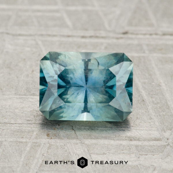 1.58-Carat Teal Montana Sapphire A rectangular, faceted 1.58-carat Montana Sapphire in blue-green rests on a textured gray surface with the "EARTH'S TREASURY" logo at the bottom.