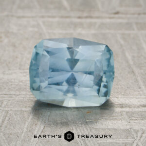 A 2.06-carat aqua blue to periwinkle unheated Montana sapphire rests on a textured metallic surface, with the "EARTH'S TREASURY" logo and text beneath it.