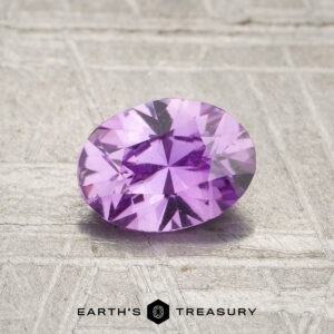 A 0.91-carat pinkish purple Ceylon sapphire (heated), oval-cut and faceted, is displayed on a textured surface with the "EARTH'S TREASURY" logo and text at the bottom.