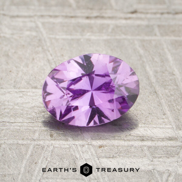 0.91-Carat Pinkish Purple Ceylon Sapphire (Heated) A 0.91-carat pinkish purple Ceylon sapphire (heated), oval-cut and faceted, is displayed on a textured surface with the "EARTH'S TREASURY" logo and text at the bottom.