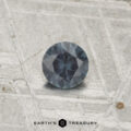 A 0.29-carat color-change garnet with a dark blue-gray hue rests on a textured metallic surface, with the text "EARTH'S TREASURY" visible at the bottom.