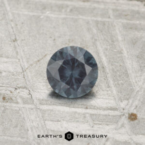 A 0.29-carat color-change garnet with a dark blue-gray hue rests on a textured metallic surface, with the text "EARTH'S TREASURY" visible at the bottom.