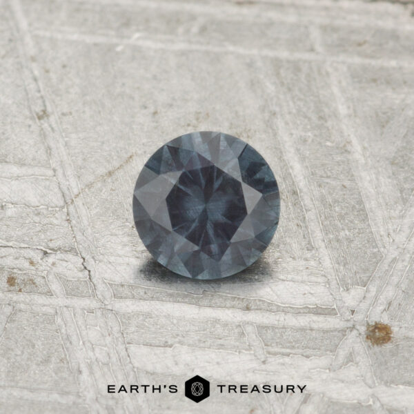 0.29-Carat Color-Change Garnet A 0.29-carat color-change garnet with a dark blue-gray hue rests on a textured metallic surface, with the text "EARTH'S TREASURY" visible at the bottom.