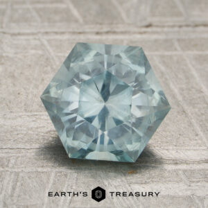 A hexagon-shaped, light blue 1.96-carat unheated Aqua Montana sapphire displayed on a textured gray surface, with "EARTH'S TREASURY" and a hexagon logo below.