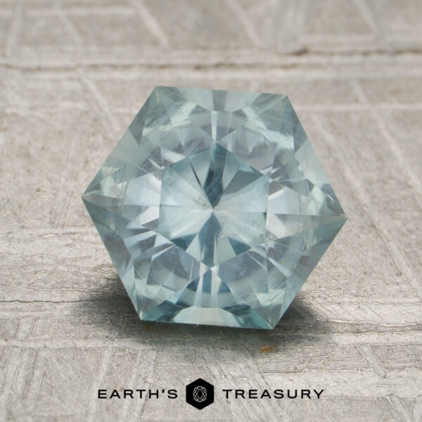1.96-Carat Aqua Montana Sapphire (Unheated) A hexagon-shaped, light blue 1.96-carat unheated Aqua Montana sapphire displayed on a textured gray surface, with "EARTH'S TREASURY" and a hexagon logo below.