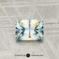 A 0.91-carat Montana Sapphire, cut in a rectangular faceted shape with blue and yellow hues, rests on a textured gray surface. "EARTH'S TREASURY" is shown at the bottom.