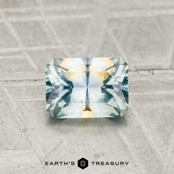 0.91-Carat Aqua-Yellow Particolored Montana Sapphire A 0.91-carat Montana Sapphire, cut in a rectangular faceted shape with blue and yellow hues, rests on a textured gray surface. "EARTH'S TREASURY" is shown at the bottom.