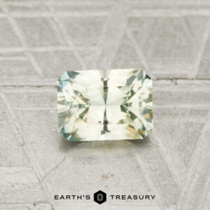 A 0.88-carat Montana sapphire with rectangular facets and light green-yellow hues rests on a textured gray surface, with the "Earth’s Treasury" logo and text below.