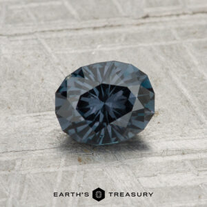 A 0.96-carat color-change garnet, round and faceted, is displayed on a gray textured surface with "EARTH'S TREASURY" at the bottom.