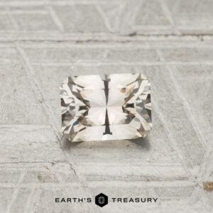 A 0.69-carat Montana Sapphire with a rectangular faceted cut and light, transparent appearance rests on a textured metallic surface; the "Earth's Treasury" logo is displayed at the bottom.