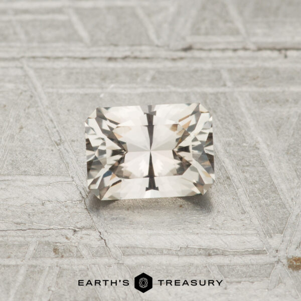 0.69-Carat Montana Sapphire A 0.69-carat Montana Sapphire with a rectangular faceted cut and light, transparent appearance rests on a textured metallic surface; the "Earth's Treasury" logo is displayed at the bottom.
