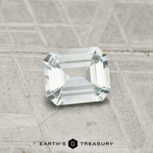 A 0.62-carat Montana sapphire with an emerald cut is displayed on a textured gray surface, with "EARTH'S TREASURY" text at the bottom.