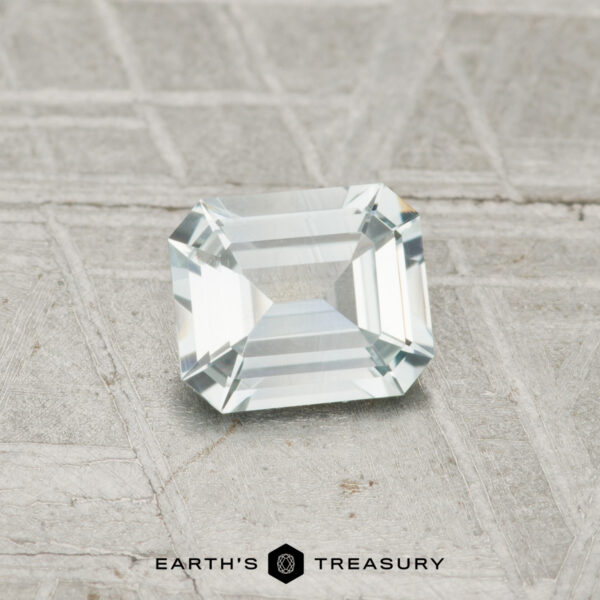 0.62-Carat Near-Colorless Montana Sapphire A 0.62-carat Montana sapphire with an emerald cut is displayed on a textured gray surface, with "EARTH'S TREASURY" text at the bottom.
