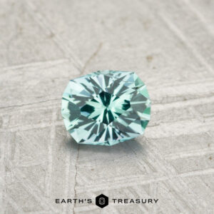 A 0.44-carat Kornerupine gemstone in mint green rests on a textured gray surface, with the text "EARTH'S TREASURY" displayed at the bottom.