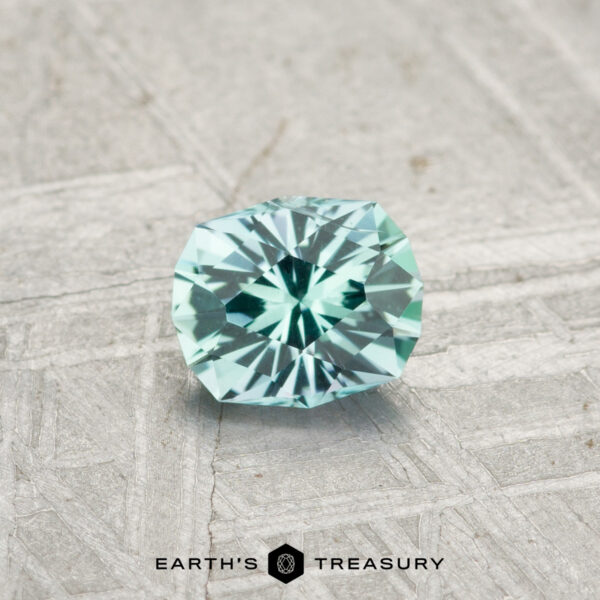 0.44-Carat Kornerupine A 0.44-carat Kornerupine gemstone in mint green rests on a textured gray surface, with the text "EARTH'S TREASURY" displayed at the bottom.