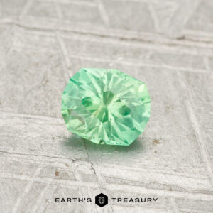 A 0.43-carat, round brilliant-cut green Kornerupine gemstone rests on a light, textured surface with "EARTH'S TREASURY" at the bottom.