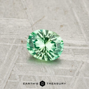A round, faceted 0.37-carat Kornerupine gemstone is shown on a textured gray surface with "Earth's Treasury" below.