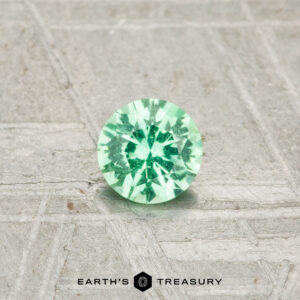 A round, faceted 0.26-carat Kornerupine gemstone rests on a textured, light surface with "EARTH'S TREASURY" displayed at the bottom.