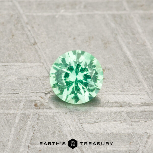 0.26-Carat Kornerupine A round, faceted 0.26-carat Kornerupine gemstone rests on a textured, light surface with "EARTH'S TREASURY" displayed at the bottom.